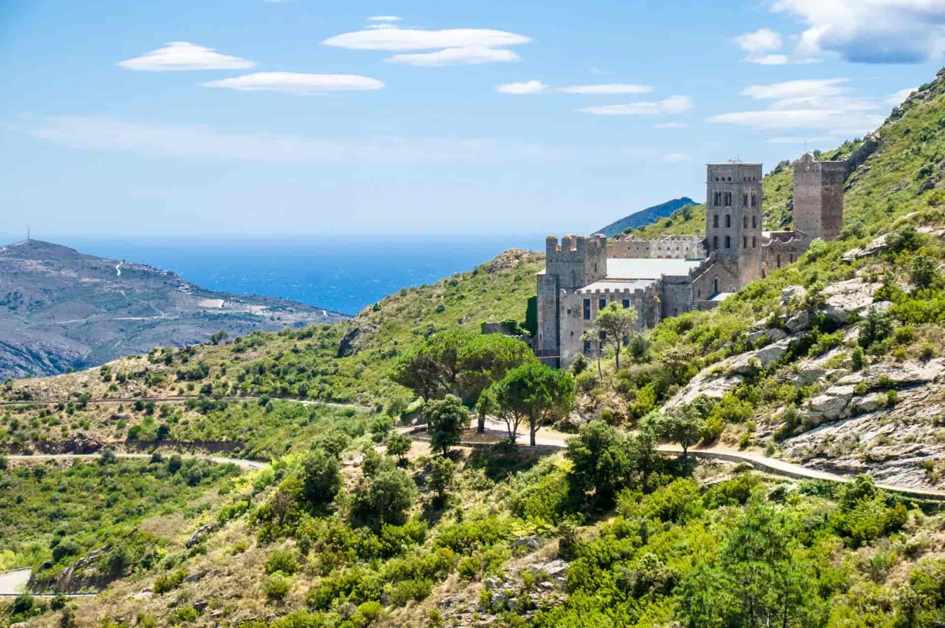Sant Pere de Rodes monastery on a green, scrub-covered hillside overlooking the blue Mediterranean Sea.