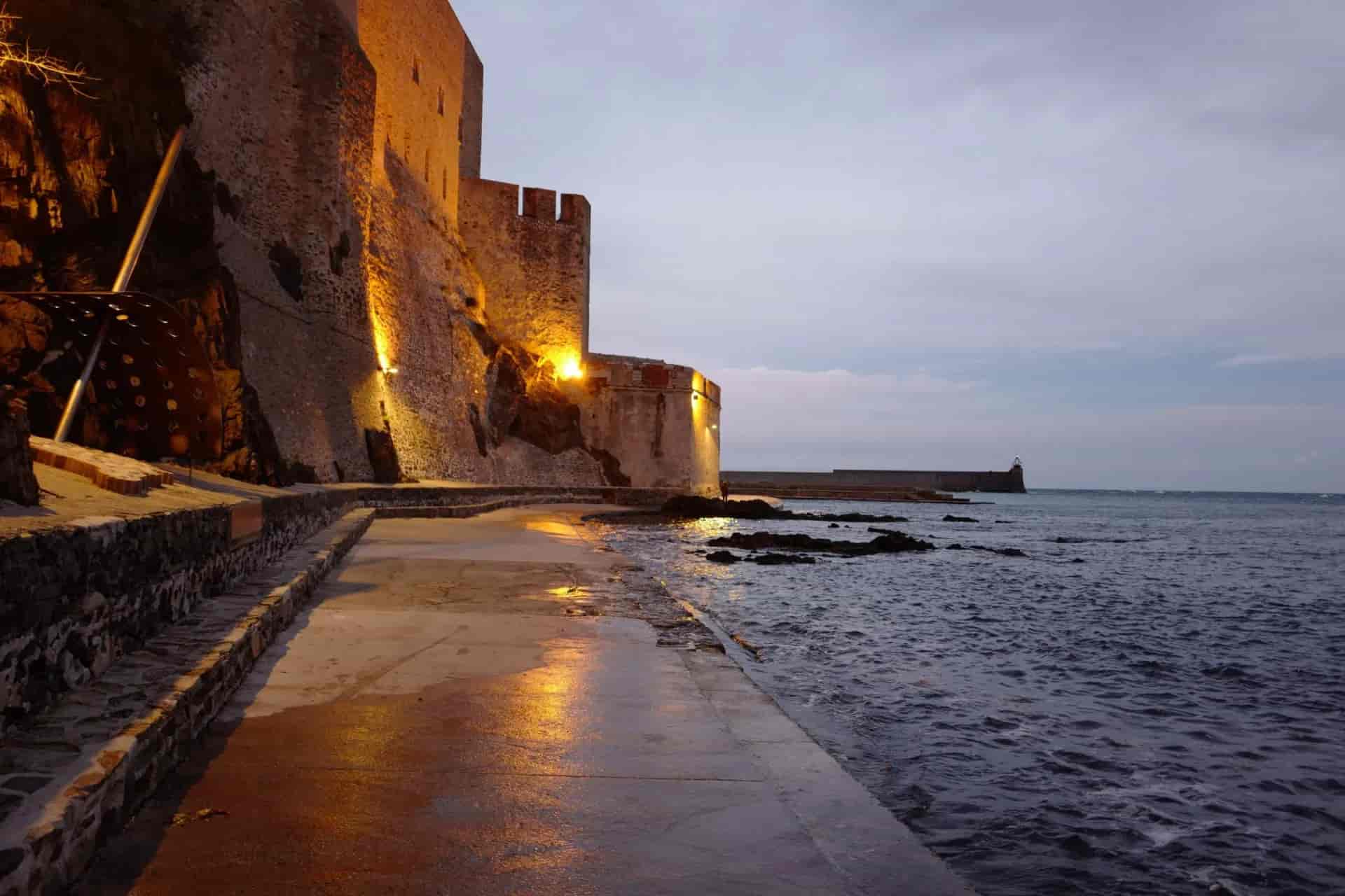 Illuminated stone fortress wall next to dark sea water at dusk in Collioure.