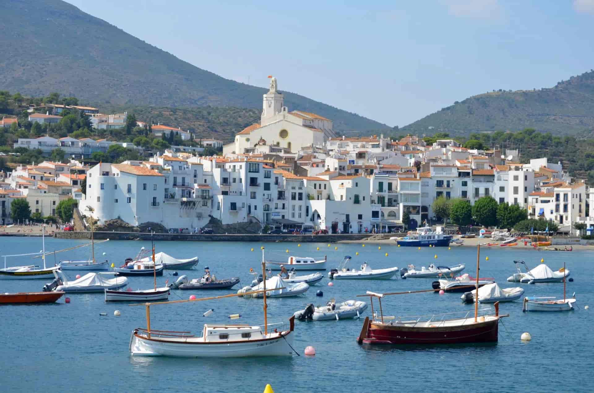 Boats anchored in blue water before white coastal town and green mountains, Cadaqués.