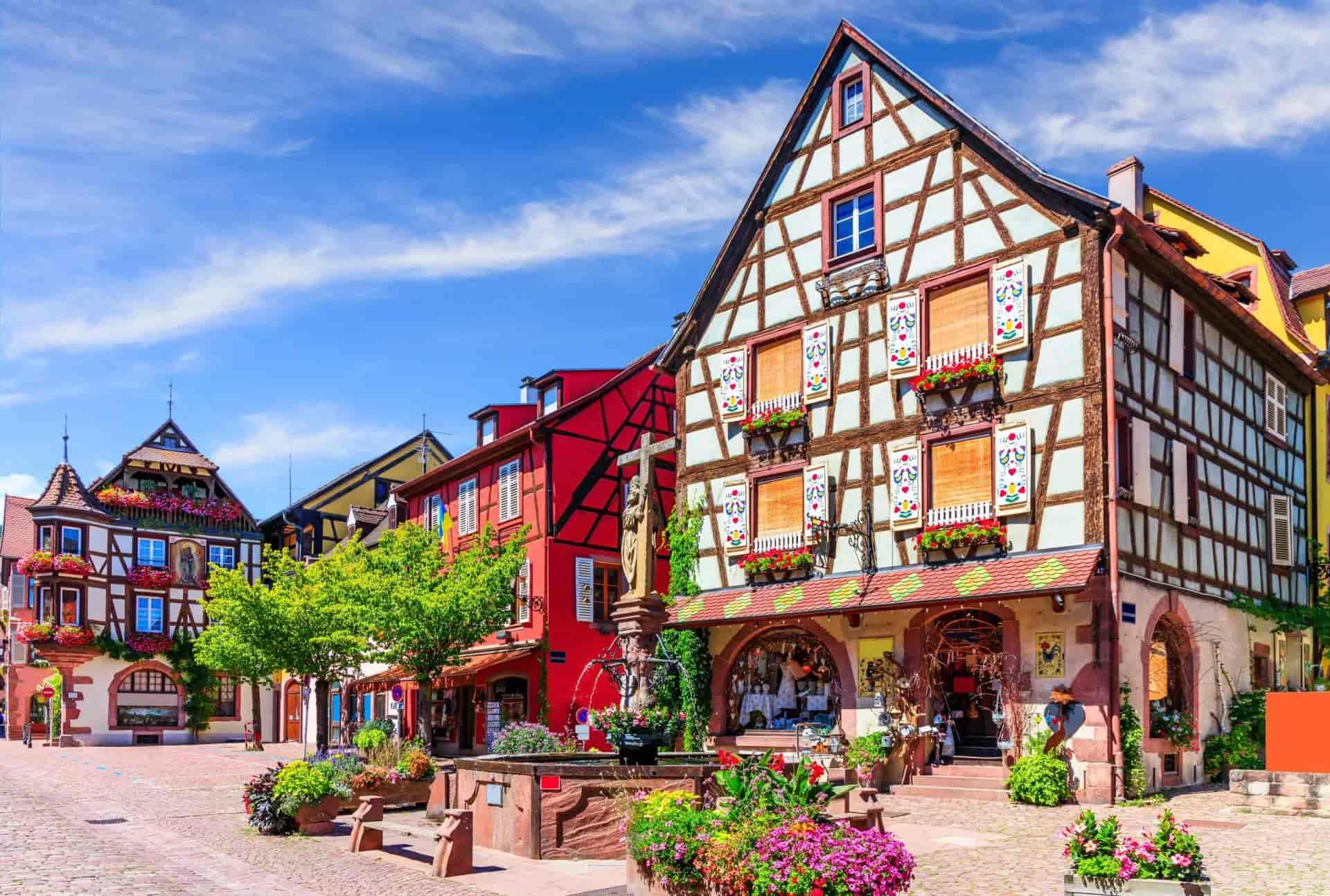 Half-timbered buildings and fountain in Kaysersberg town square with bright flowers under blue sky.