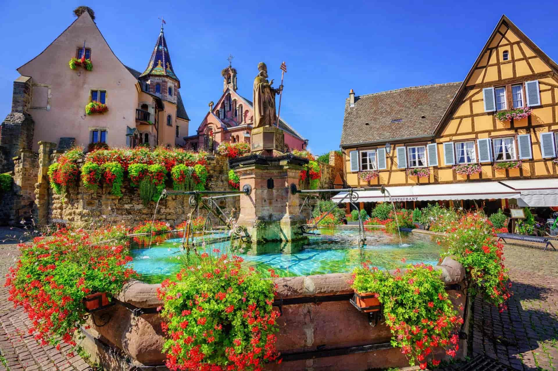 Fountain statue surrounded by red flowers in Eguisheim, France, with half-timbered buildings.