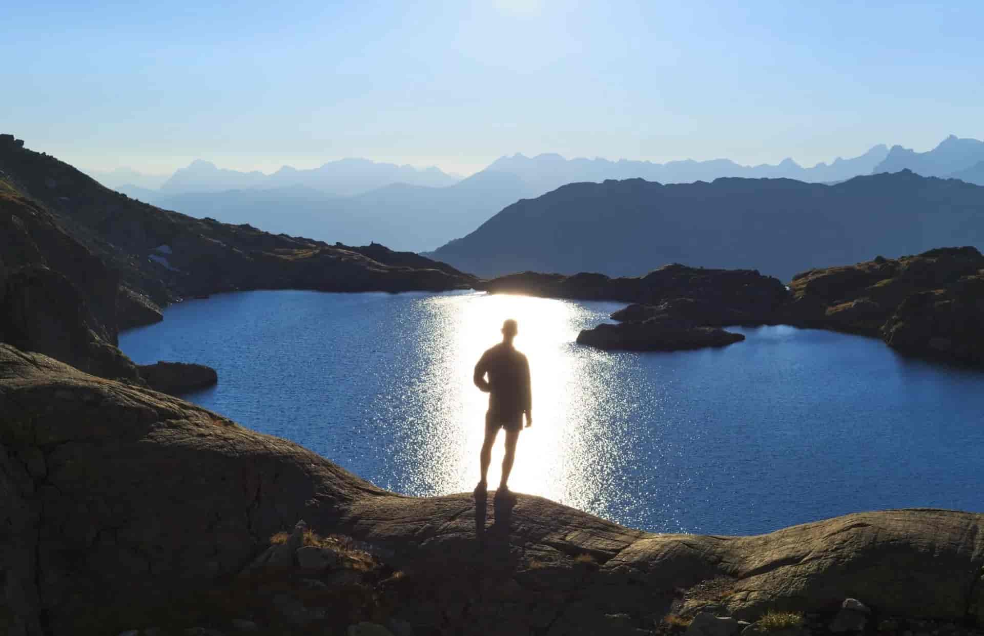 Hiker silhouetted against sunlit alpine lake with layered mountains in background