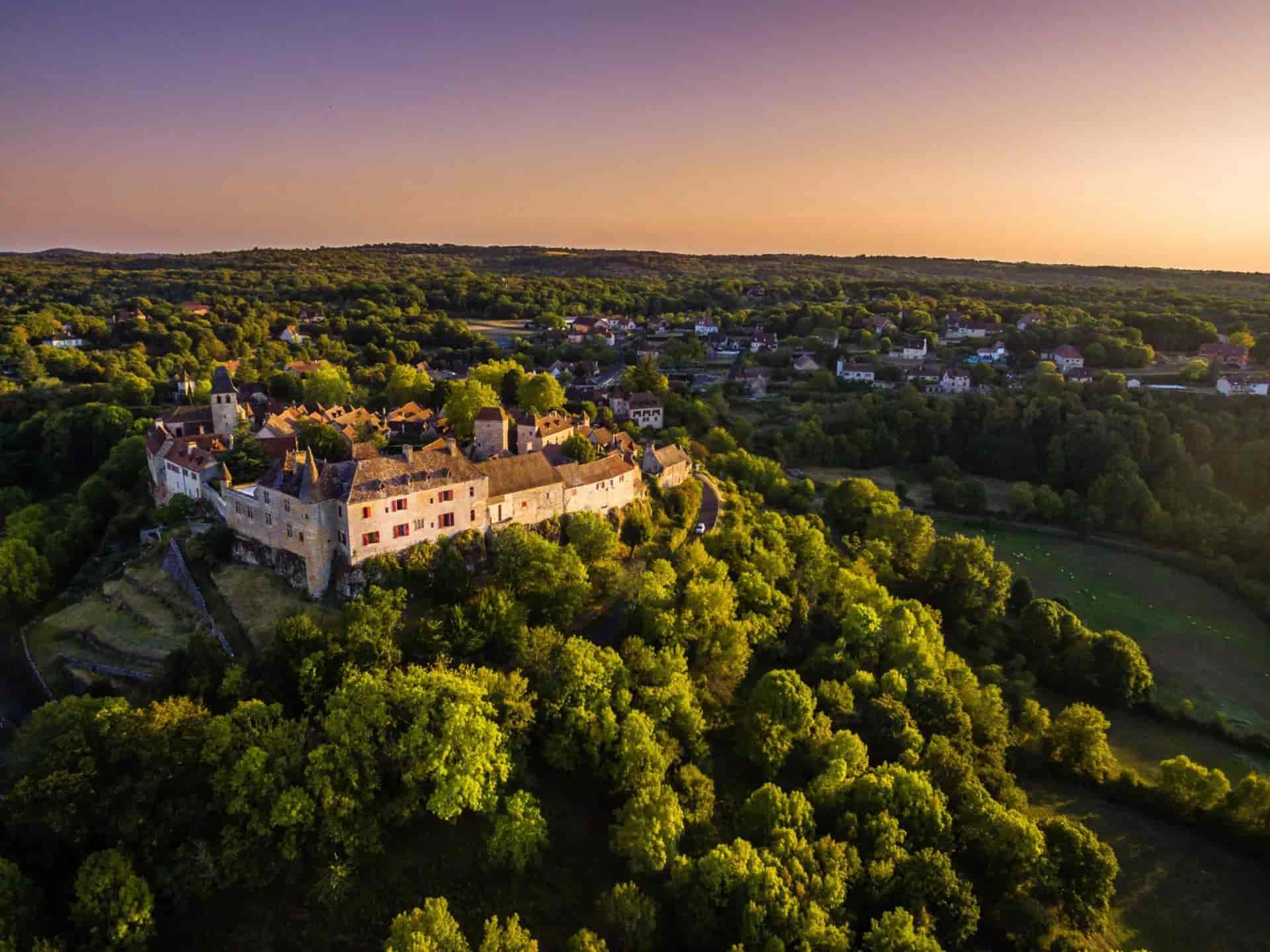 Loubressac village and chateau perched on a hill surrounded by lush green forest at sunset.