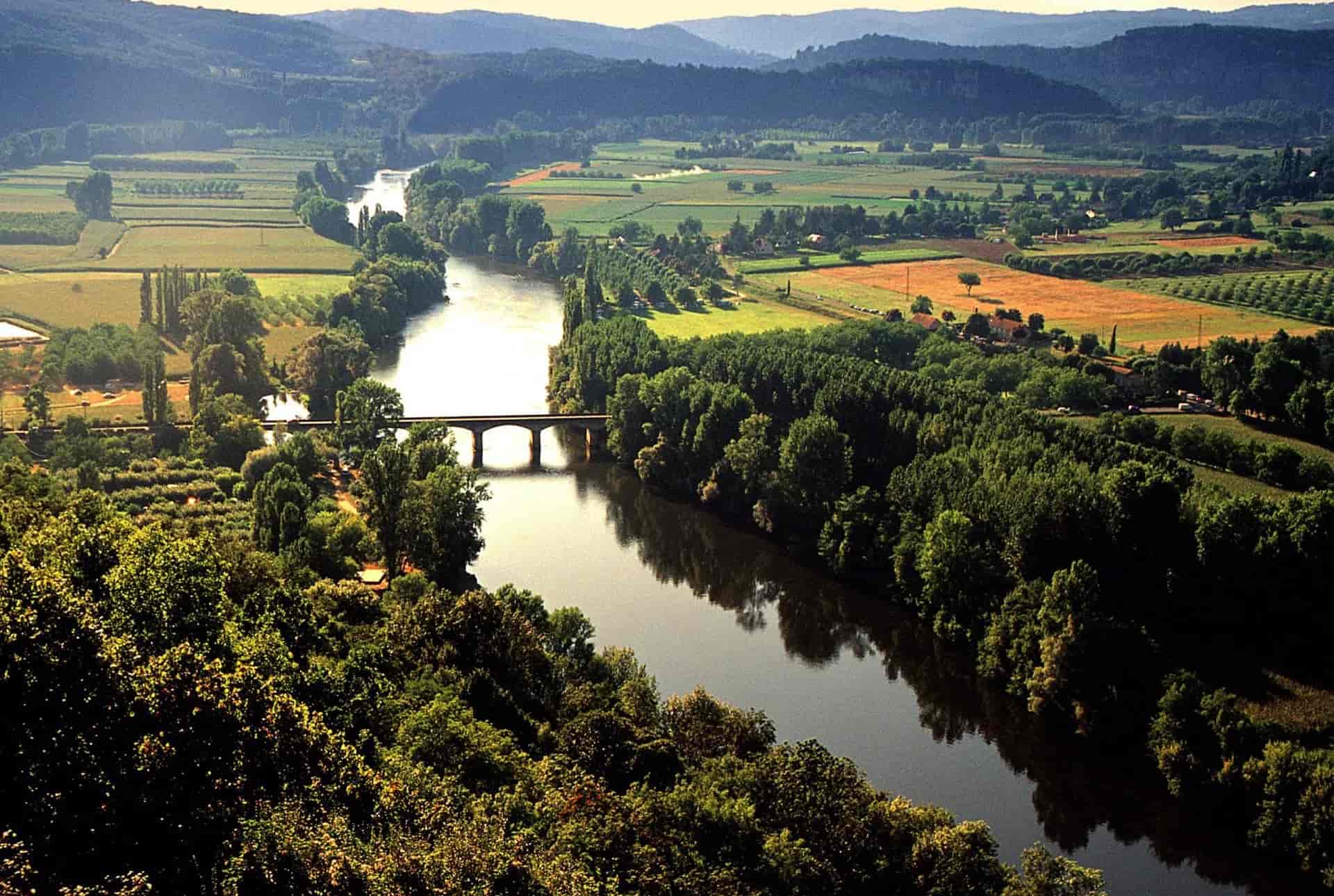 River Dordogne winding through green fields and forests with a stone bridge and rolling hills.