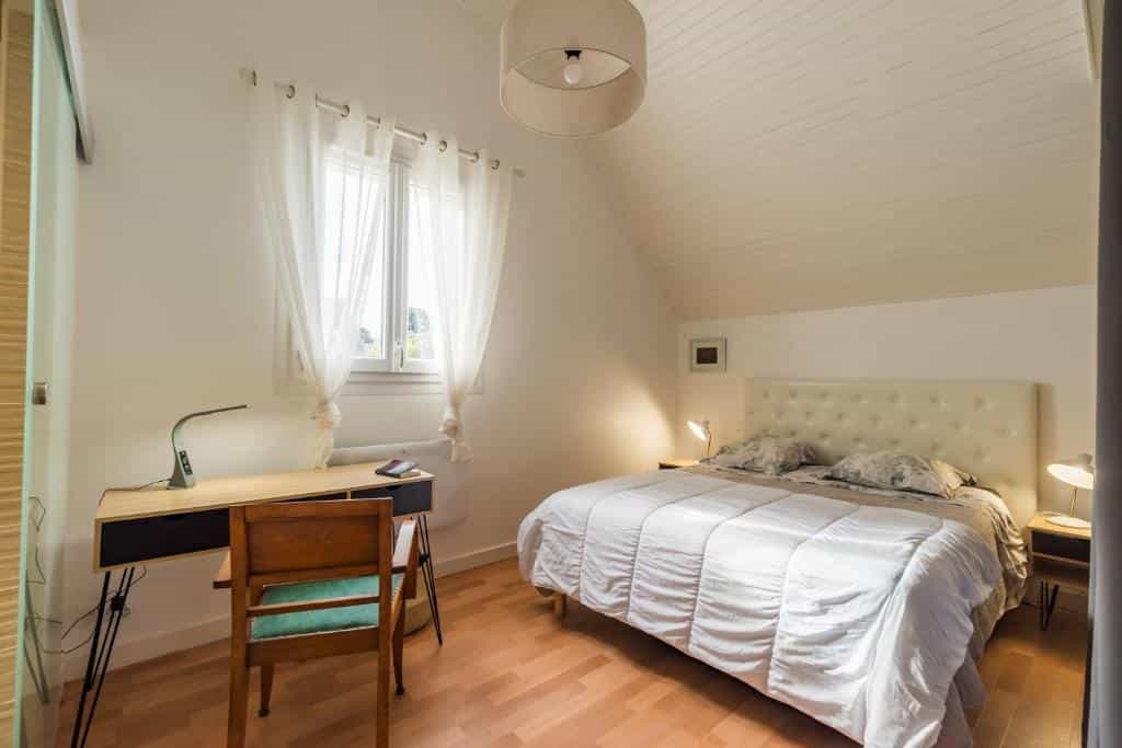 Bedroom with white walls, sloped ceiling, wood floor, desk, and large bed with white duvet.