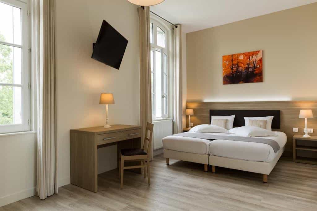 Twin beds in hotel room with desk, wall-mounted TV, and autumn landscape art