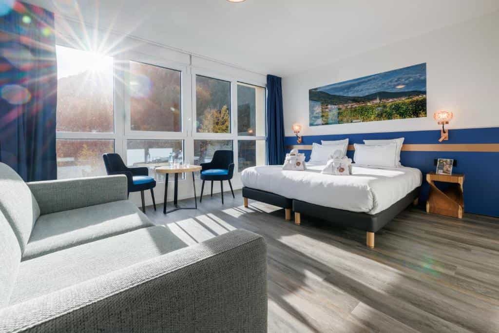 Bright hotel room with twin beds, grey sofa, and sunlit window view of trees, Husseren.