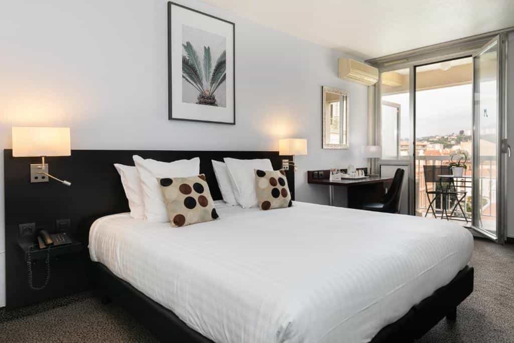 Hotel room with white bedding, balcony view of hillside town, French Riviera.