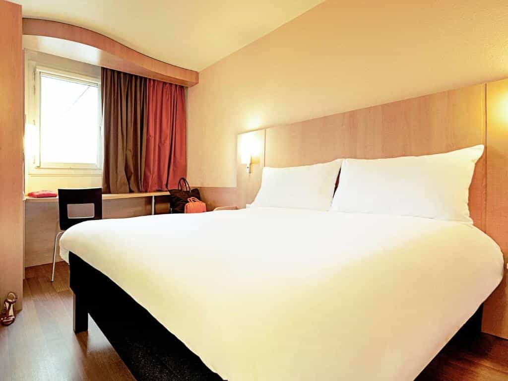 Hotel room with large white bed, wood paneling, and window with brown and red curtains.