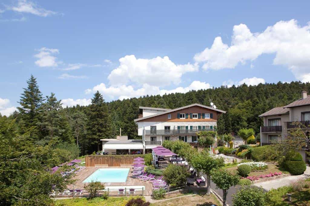 Hotel with outdoor pool and sun loungers nestled against a dense pine forest under a blue sky.