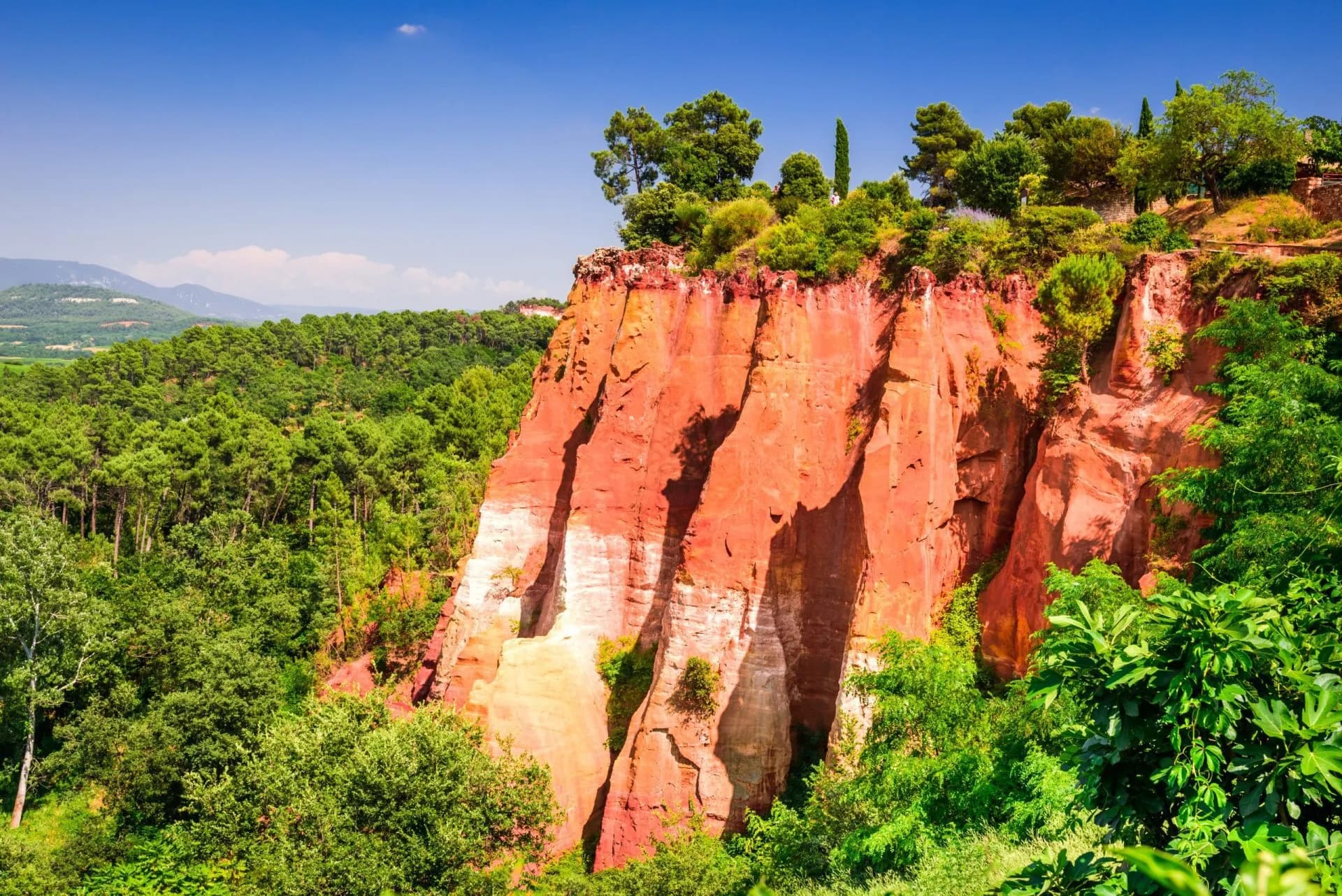 Iconic ochre cliffs in Roussillon surrounded by vibrant green trees under a clear blue sky.
