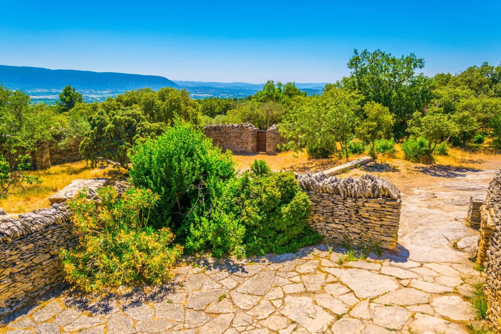 Stone walls and flagstone paths of the Village des Bories in Provence with green trees and distant hills.