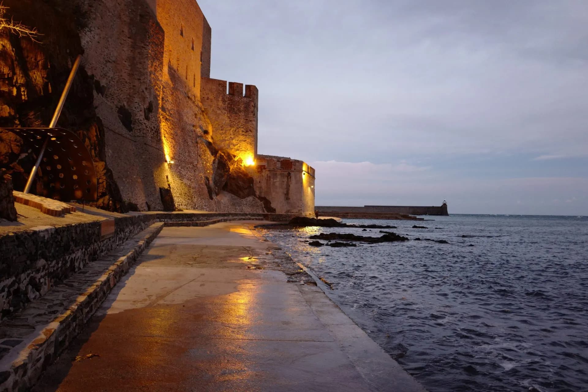 Illuminated stone fortress wall next to dark sea water at dusk in Collioure.