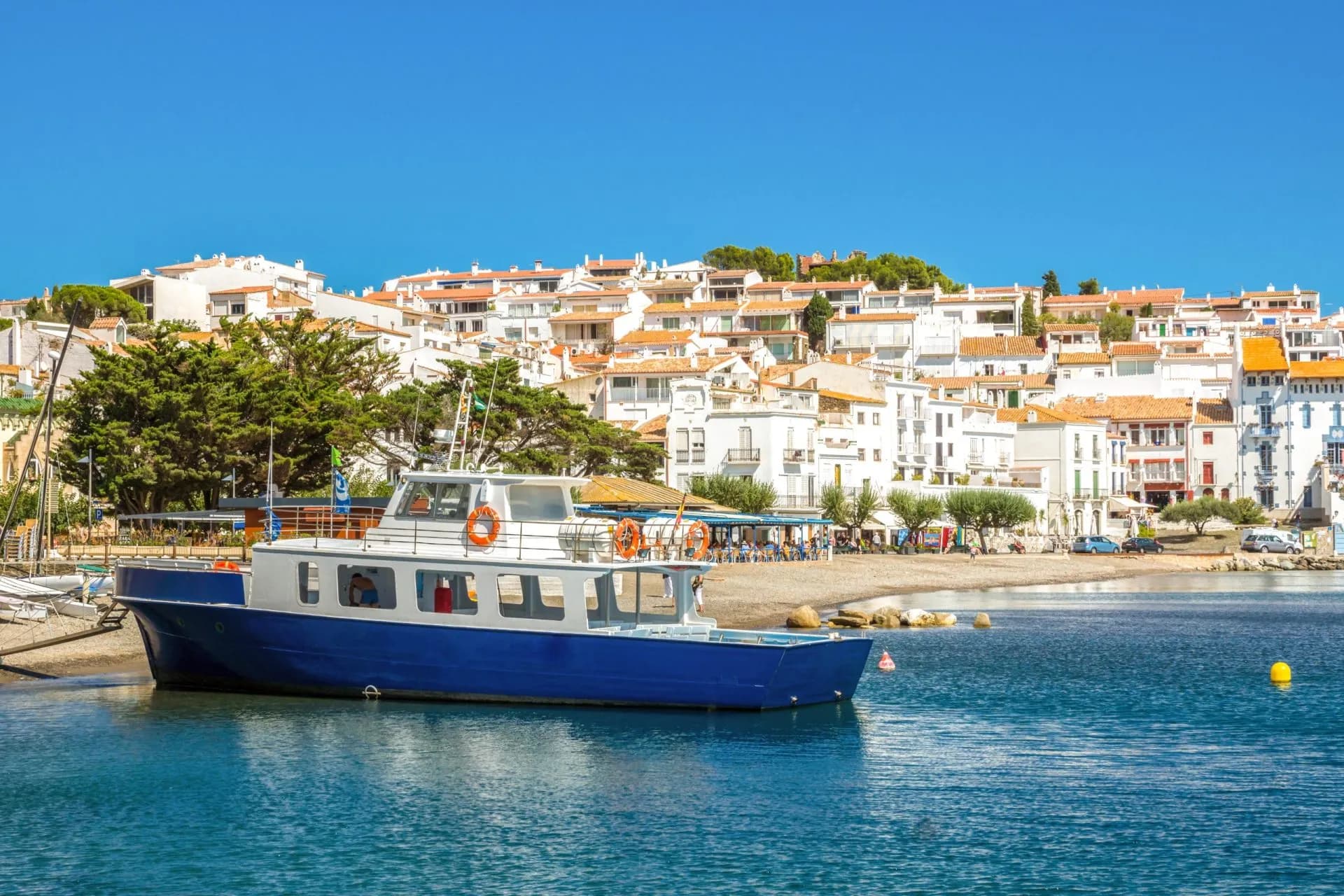 Blue and white boat in the water near the white buildings of Cadaqués coastline.