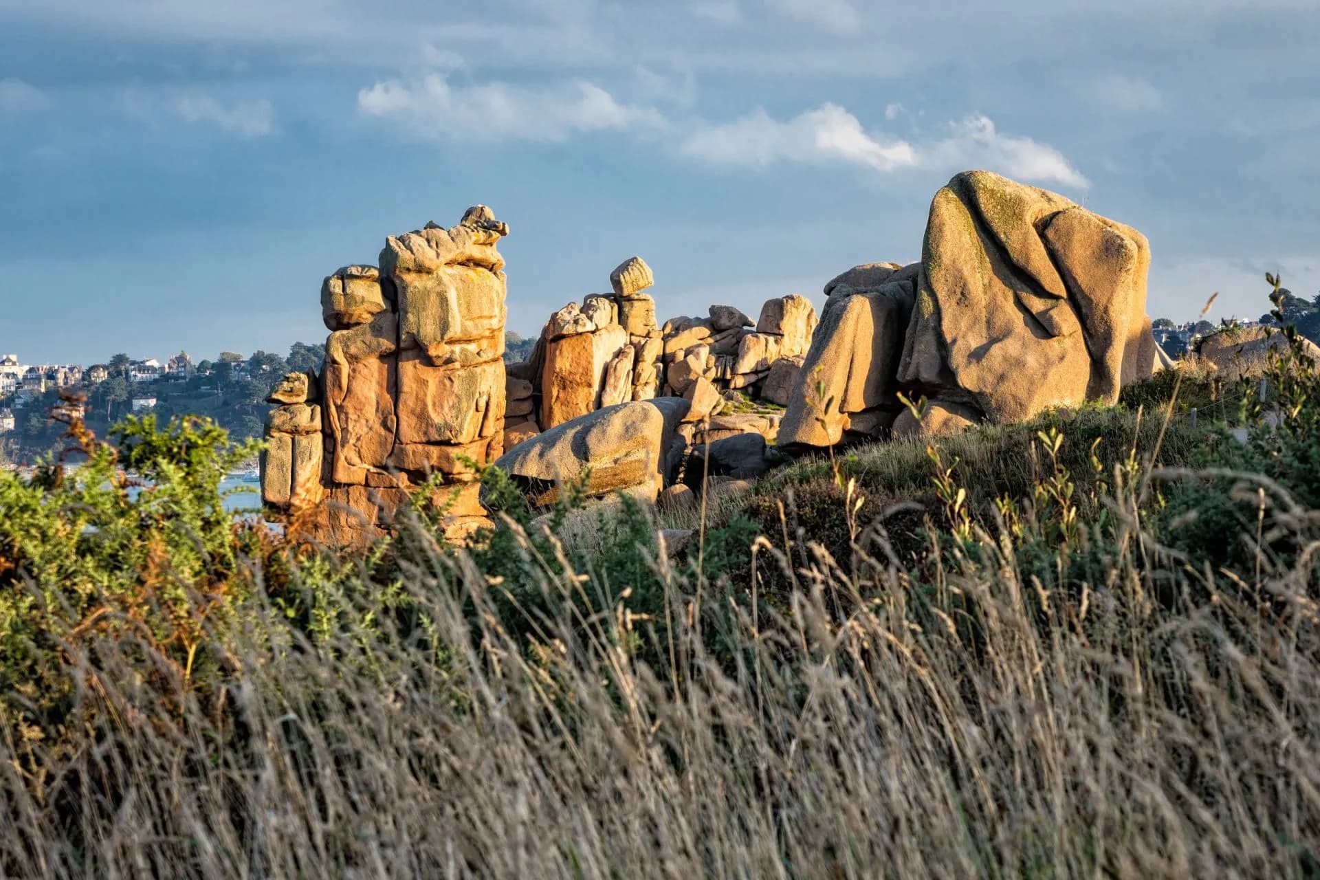 Pink granite rock formations illuminated by sunlight over coastal grasses with distant town.