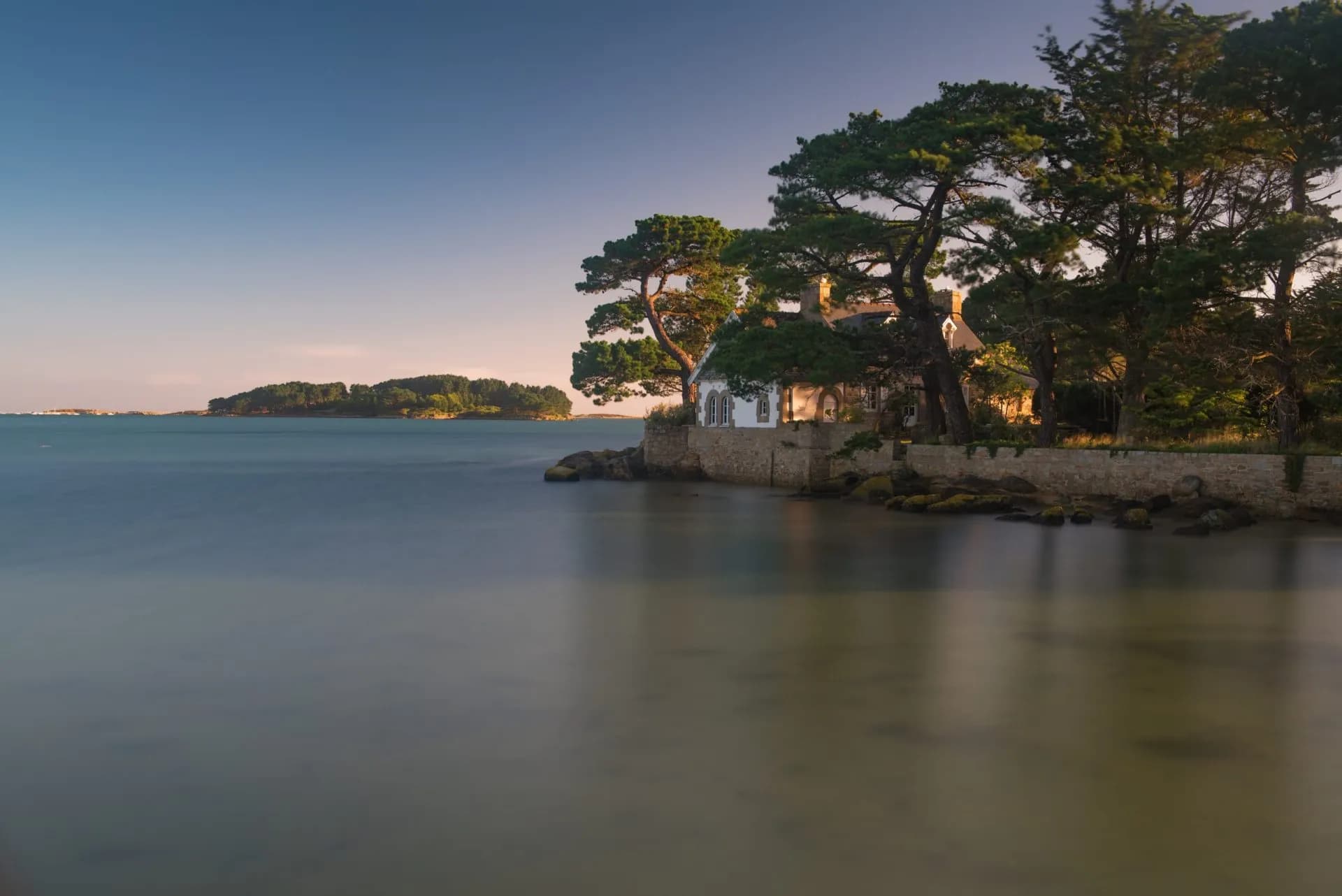 Coastal house on stone wall near Trebeurden with smooth water and wooded island in distance.