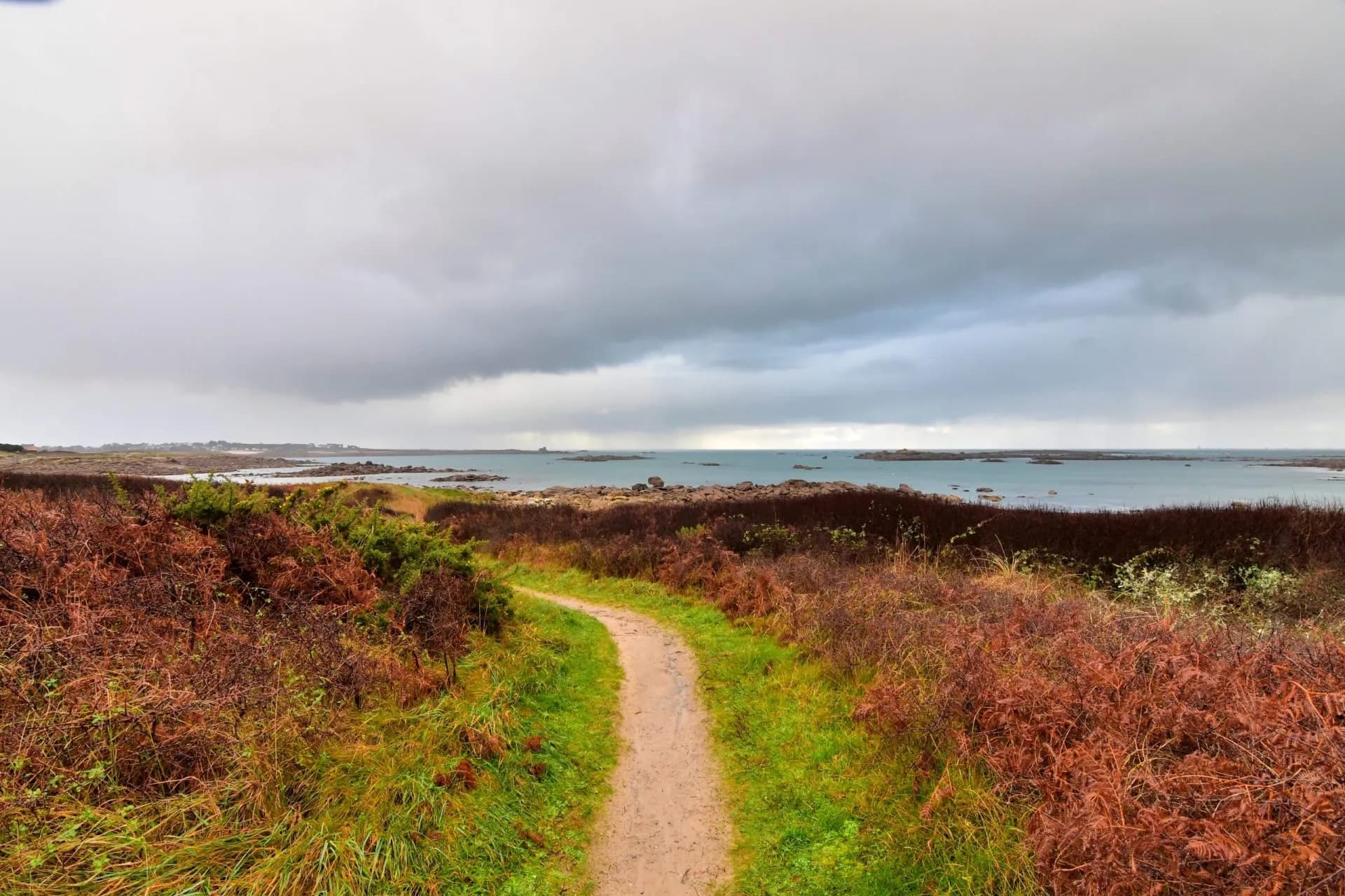 Winding dirt path through coastal scrub towards the sea under a cloudy sky, near Landrellec.