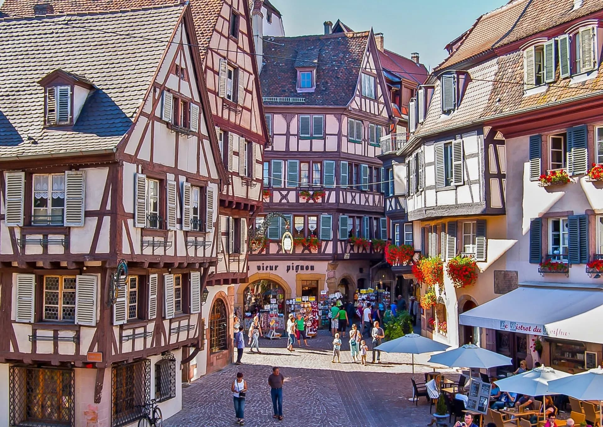 Half-timbered buildings surrounding a lively square with shops and outdoor dining in Eguisheim.