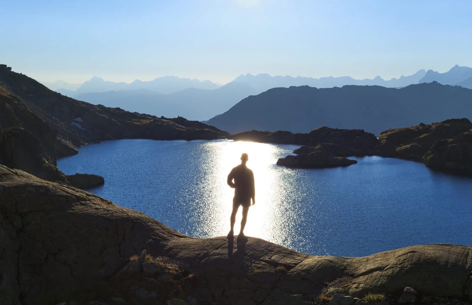 Hiker silhouetted against sunlit alpine lake with layered mountains in background