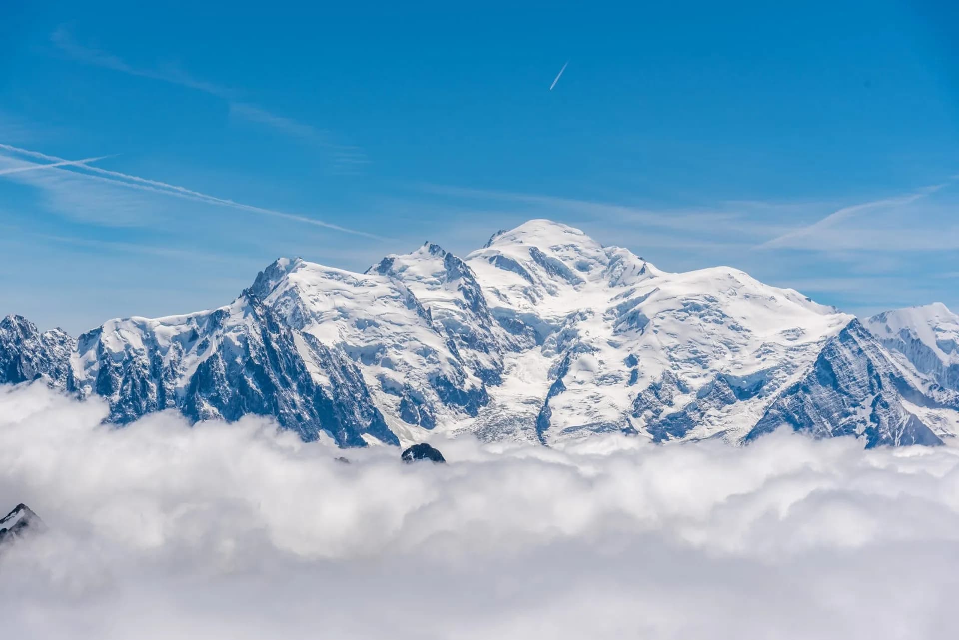 Snow-covered Mont Blanc mountain peaks rising above a sea of white clouds under a blue sky.