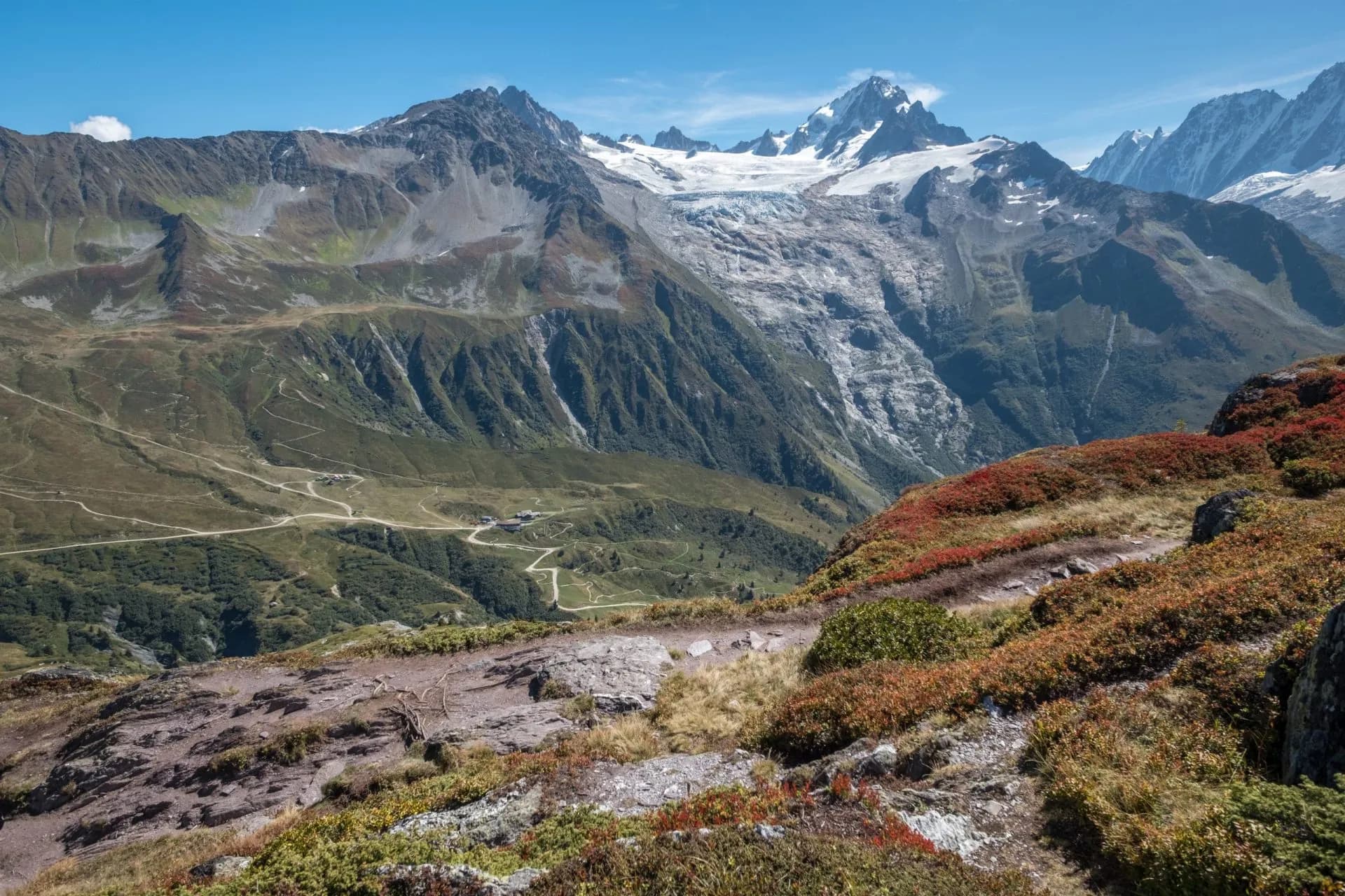 Hiking trail overlooking valley with winding roads and snow-capped alpine mountains