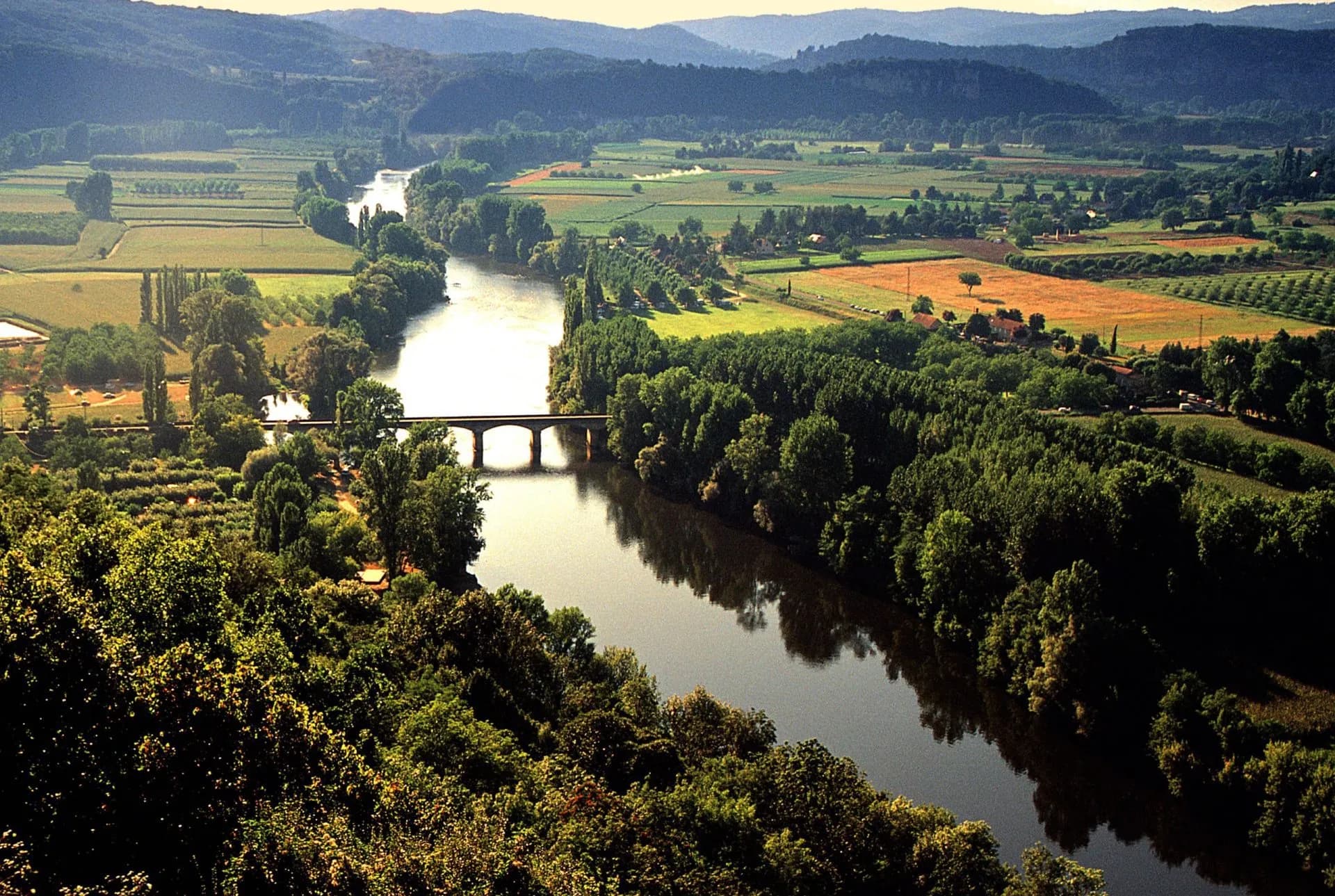 River Dordogne winding through green fields and forests with a stone bridge and rolling hills.