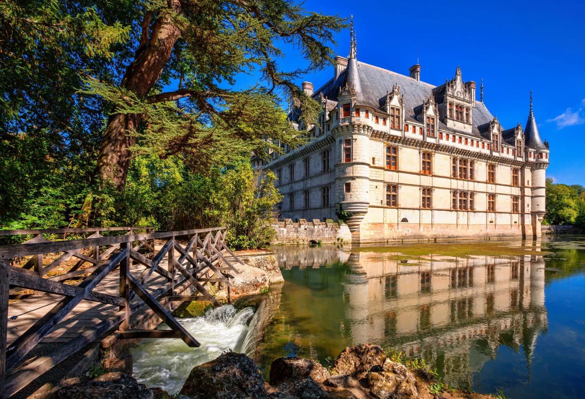 Chateau d'Azay-le-Rideau reflected in water next to a wooden bridge and waterfall.