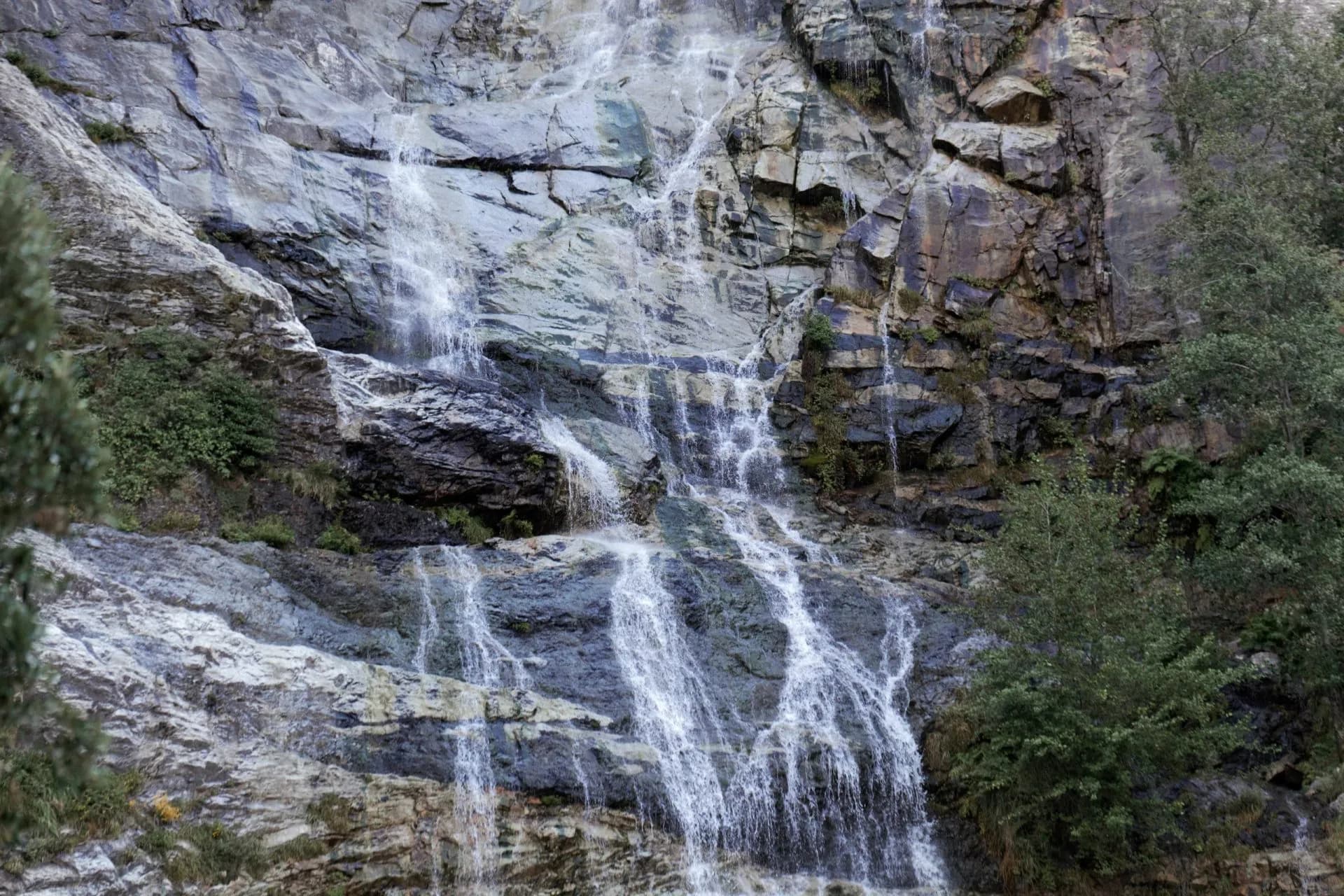 Cascading waterfall flowing down a steep, multi-toned rock face surrounded by green foliage.