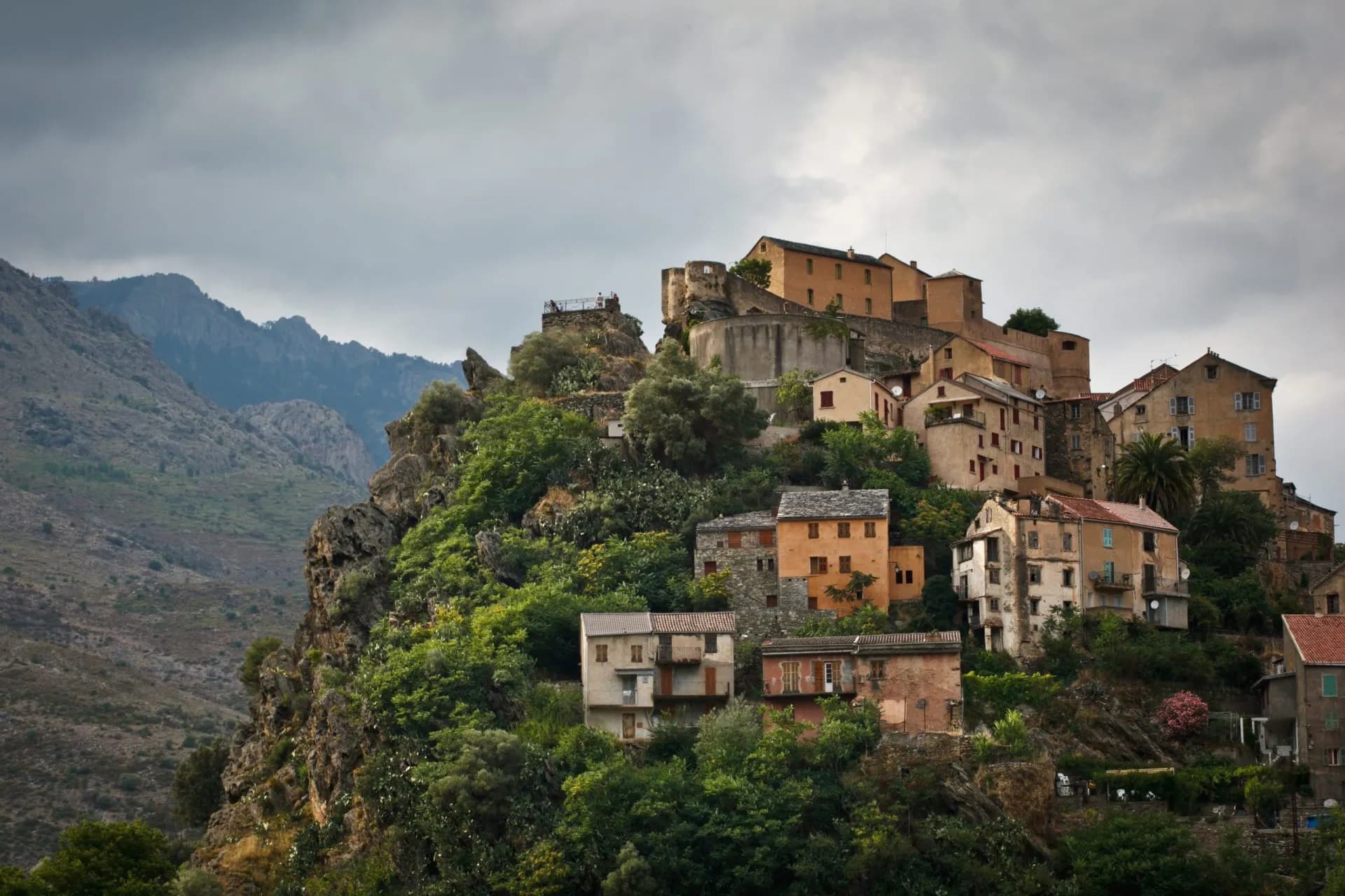 Hillside village houses built into rocky terrain with lush greenery and mountains under a cloudy sky in Corsica.