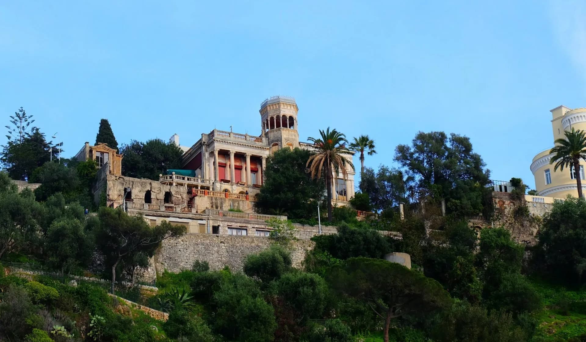 Ornate villa with a tower on a lush green hillside under a clear blue sky, Mont Boron.