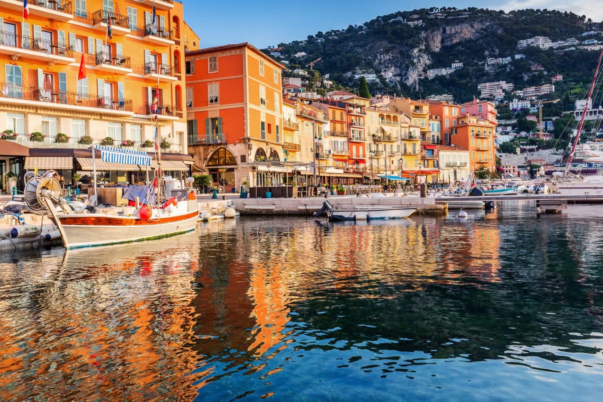Boats docked in Villefranche-sur-Mer harbor with colorful buildings and steep hillside behind.