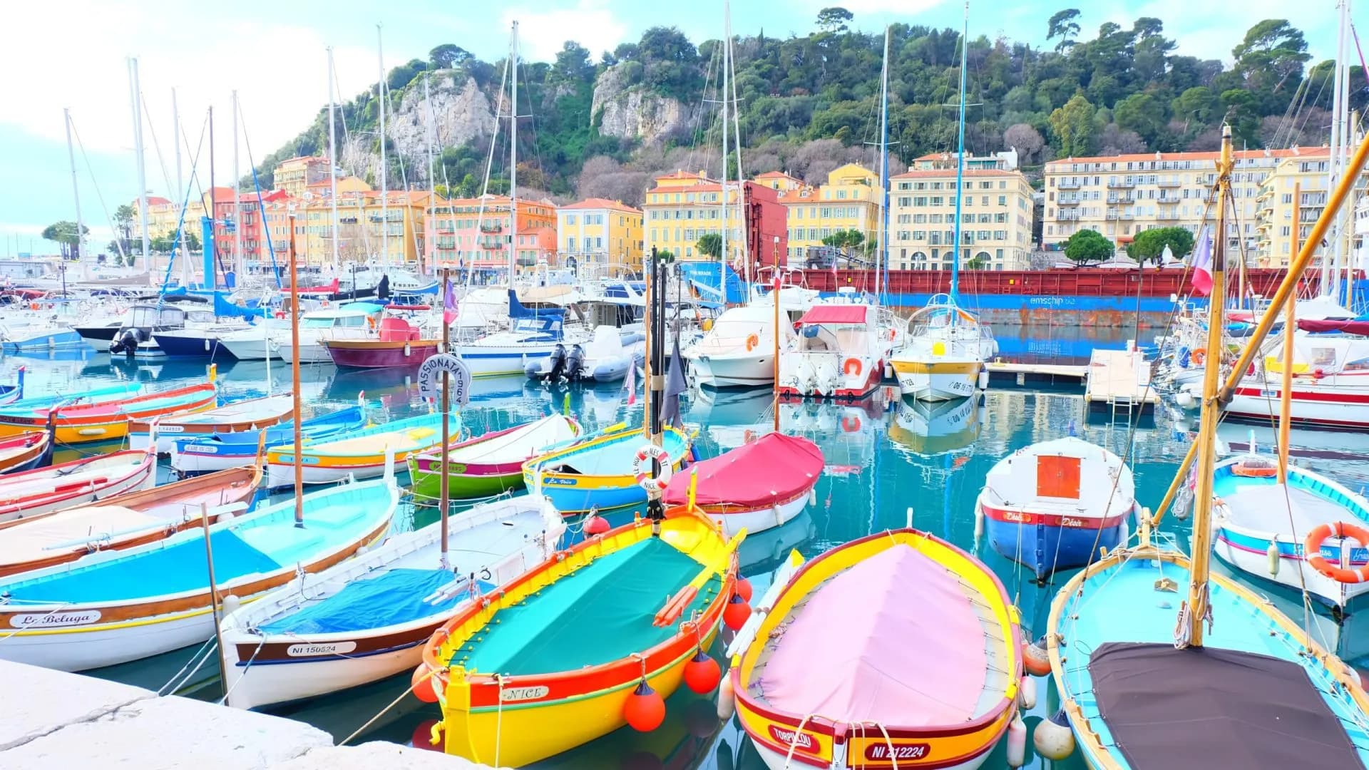 Colorful fishing boats and sailboats moored in the harbor of Nice, with colorful buildings and a hillside backdrop.