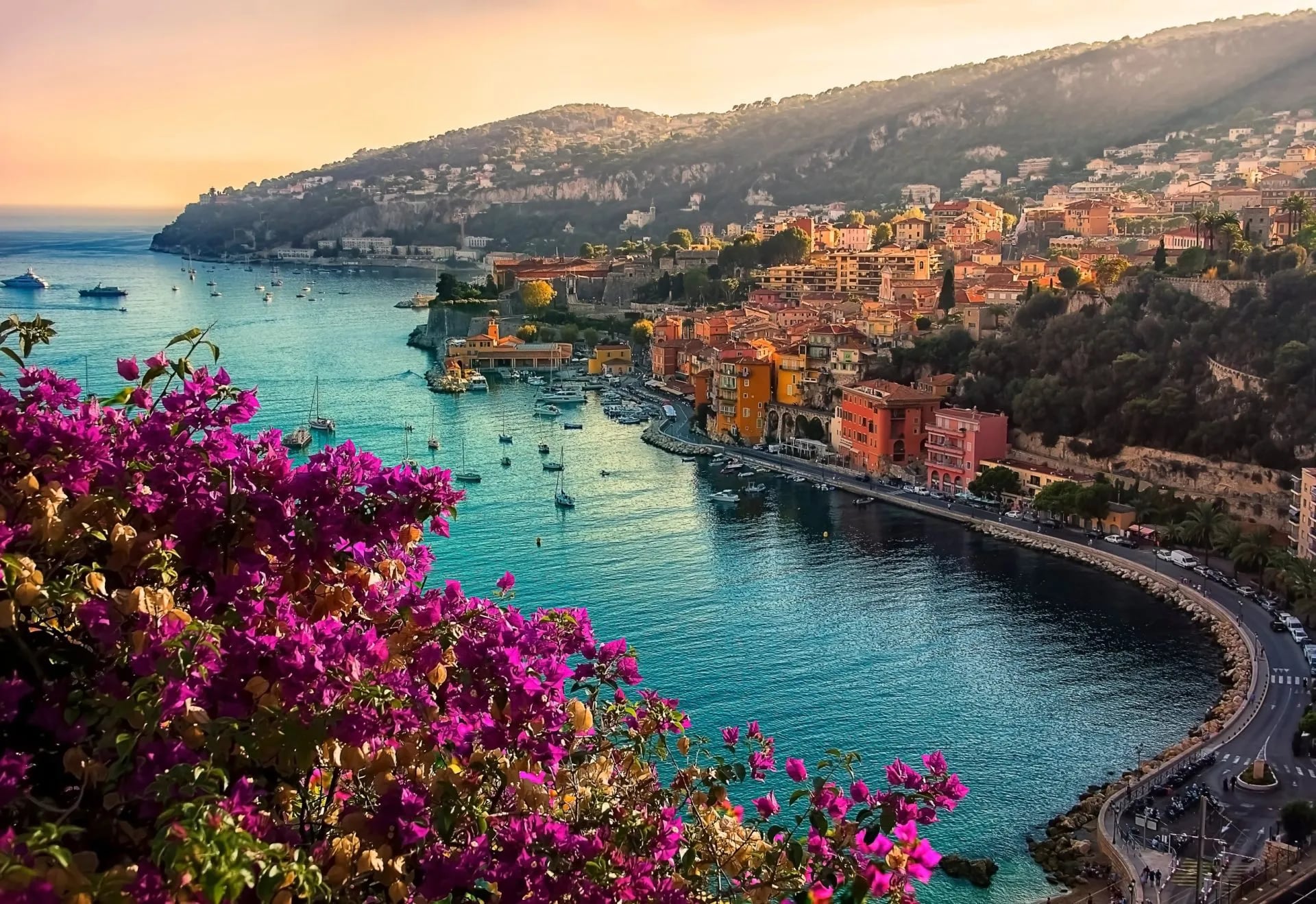 Colorful bougainvillea overlooking Villefranche-sur-Mer harbor, boats, and hillside town at sunset.