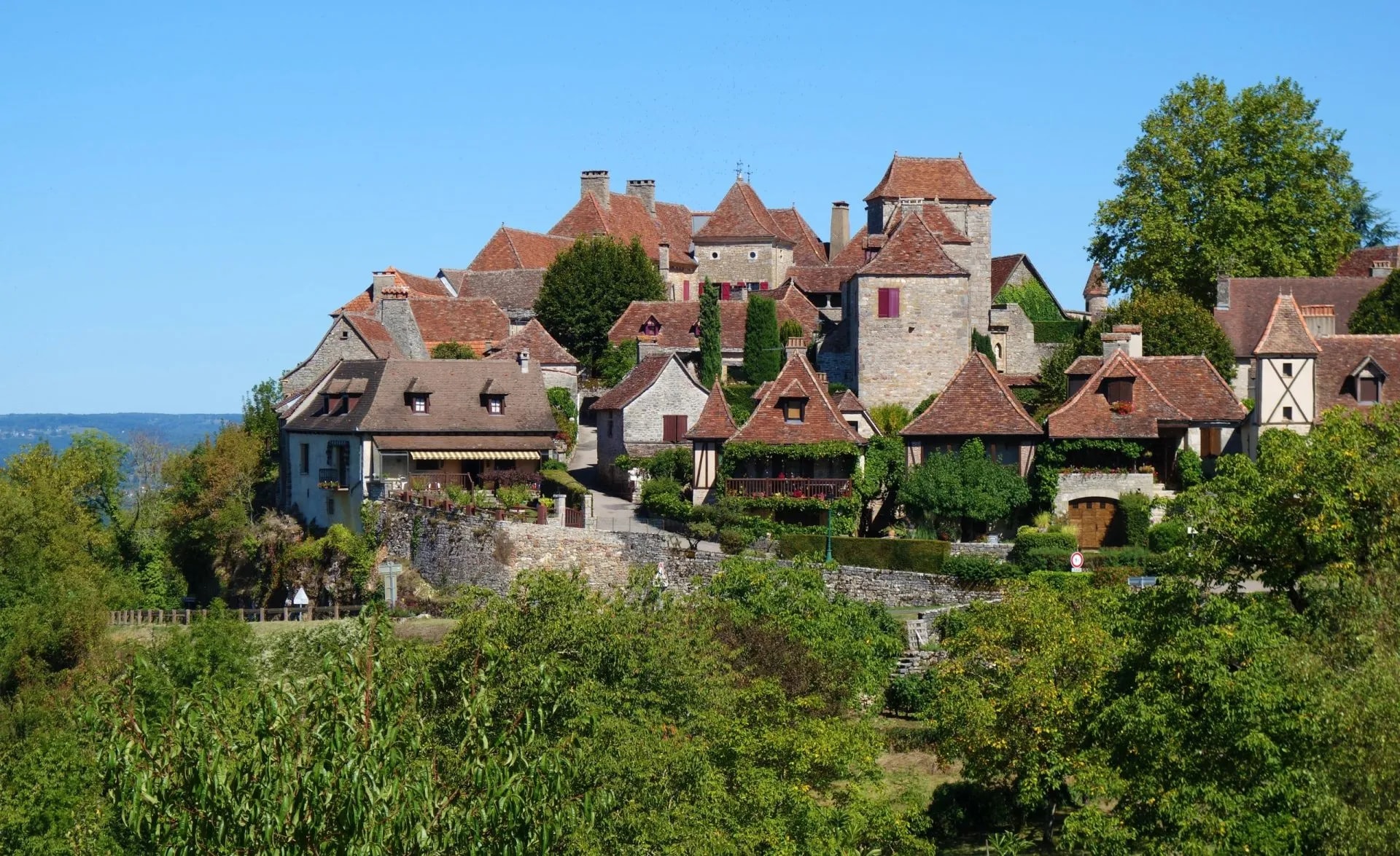 Historic stone houses with terracotta roofs clustered on a hillside in Loubressac under a clear blue sky.