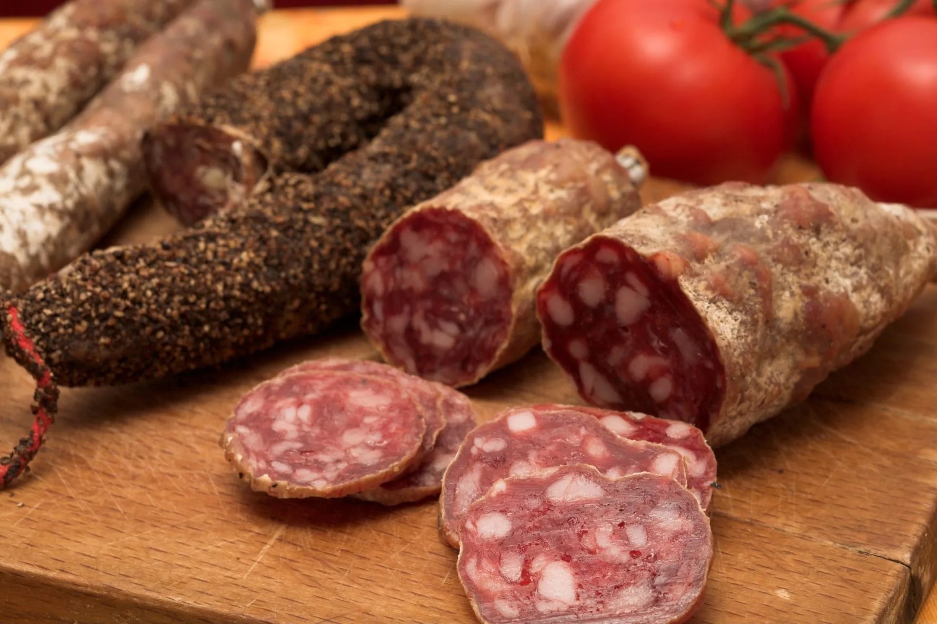 Corsican sausages, some whole and some sliced, on a wooden board with tomatoes in the background.