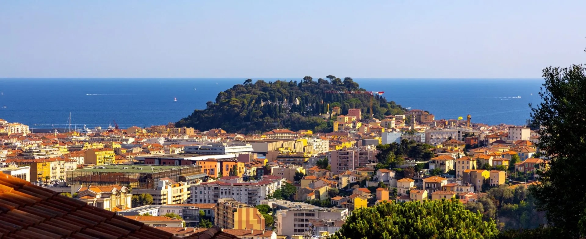 Cityscape of colorful buildings leading to a wooded hill overlooking the deep blue Mediterranean Sea.
