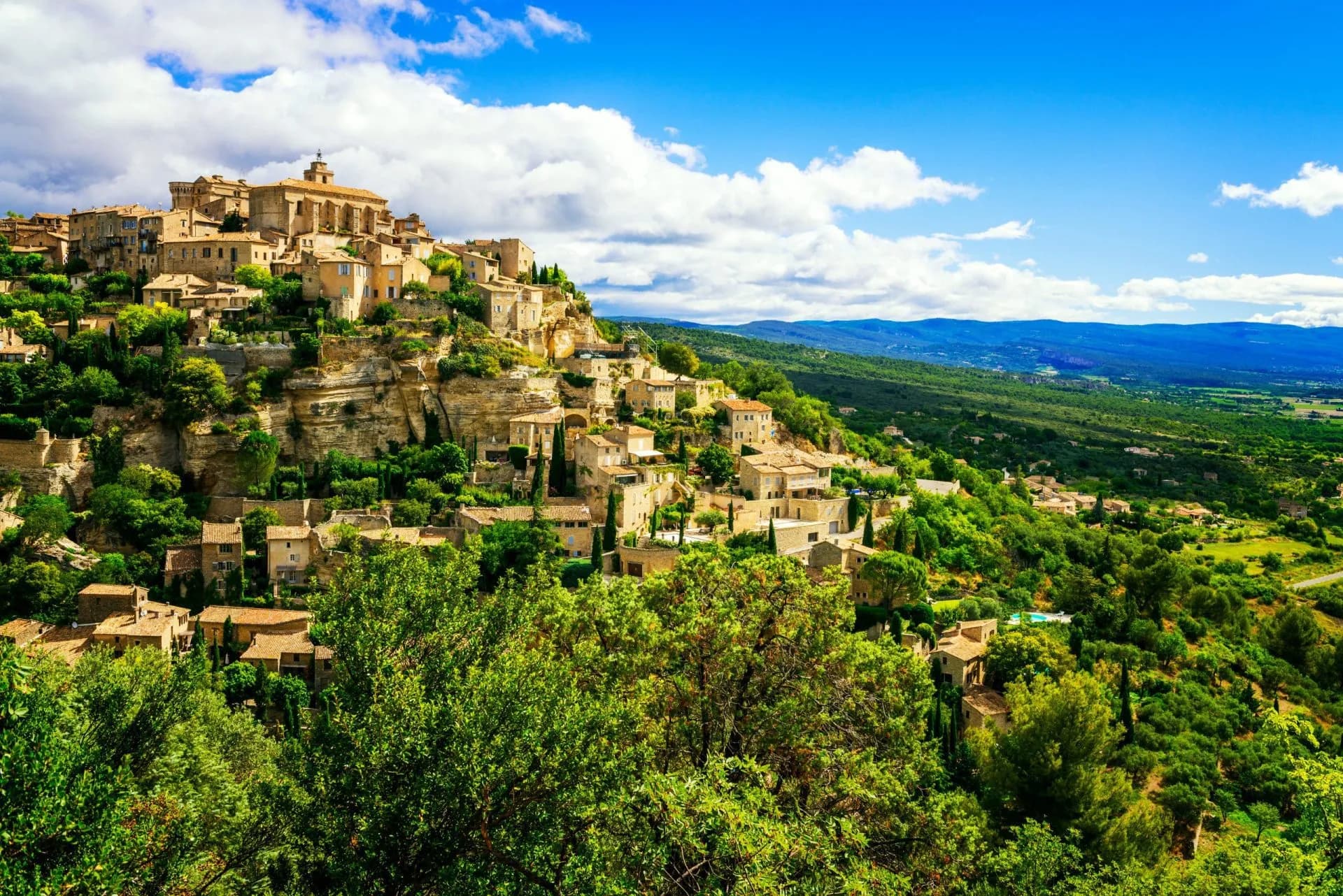 Hilltop village with stone buildings nestled in lush green landscape under a blue sky with clouds.