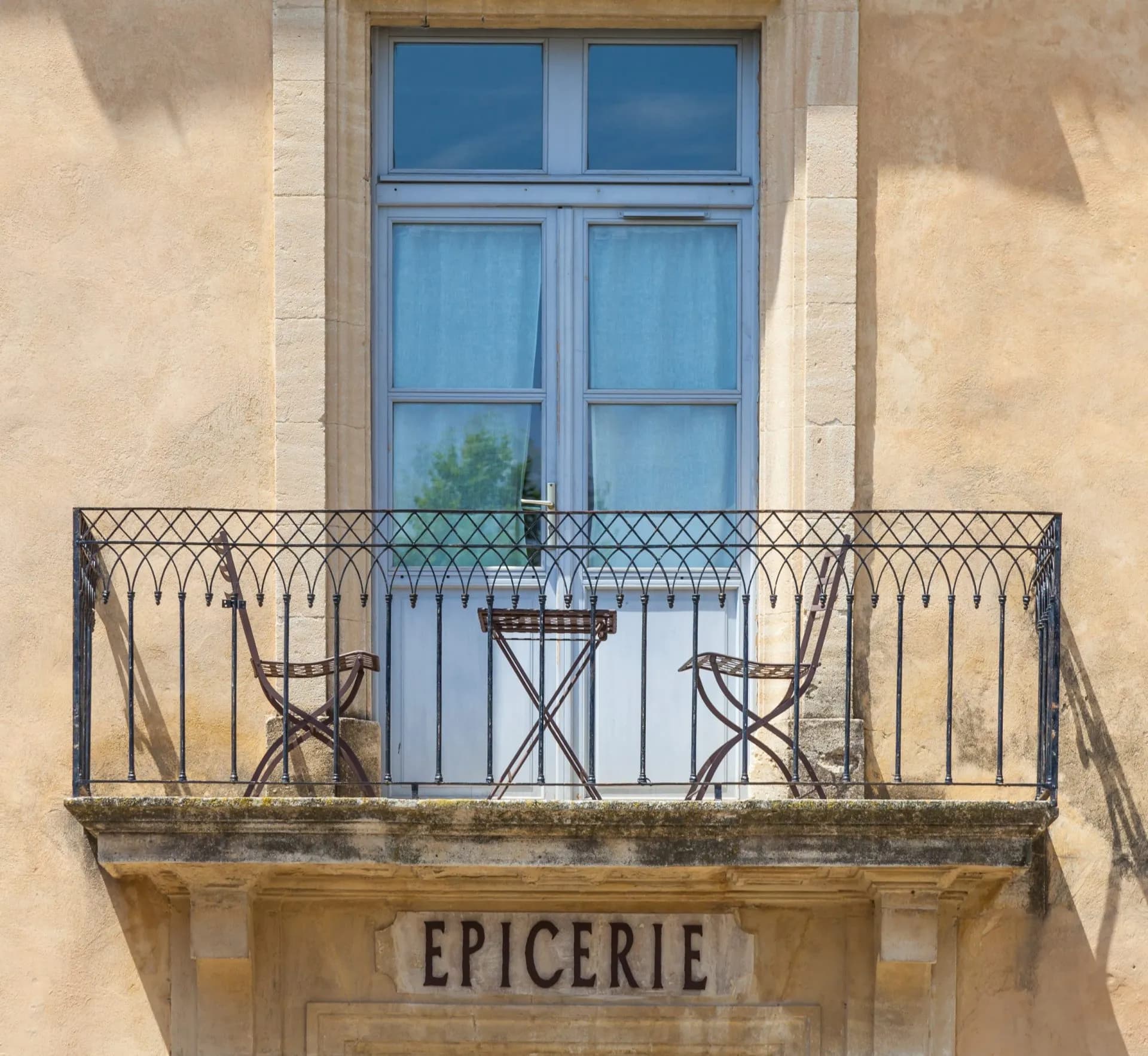 Balcony with wrought iron railing, table, and chairs above "EPICERIE" sign on stucco building.