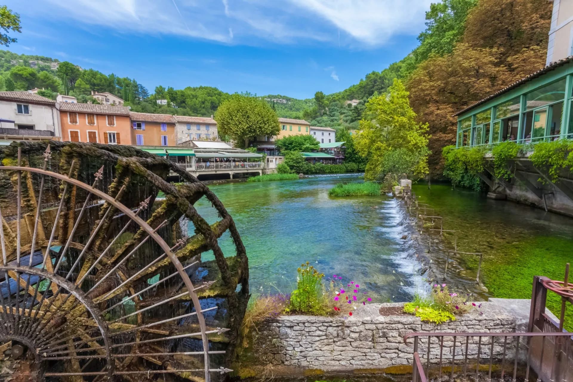 Old mossy waterwheel next to clear river in Fontaine de Vaucluse village with colorful buildings.