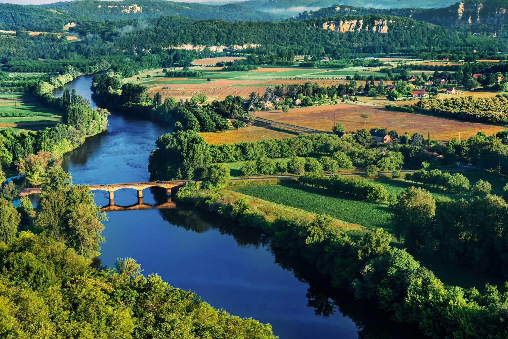 River Dordogne winding through lush green valley with stone bridge and rolling hills.