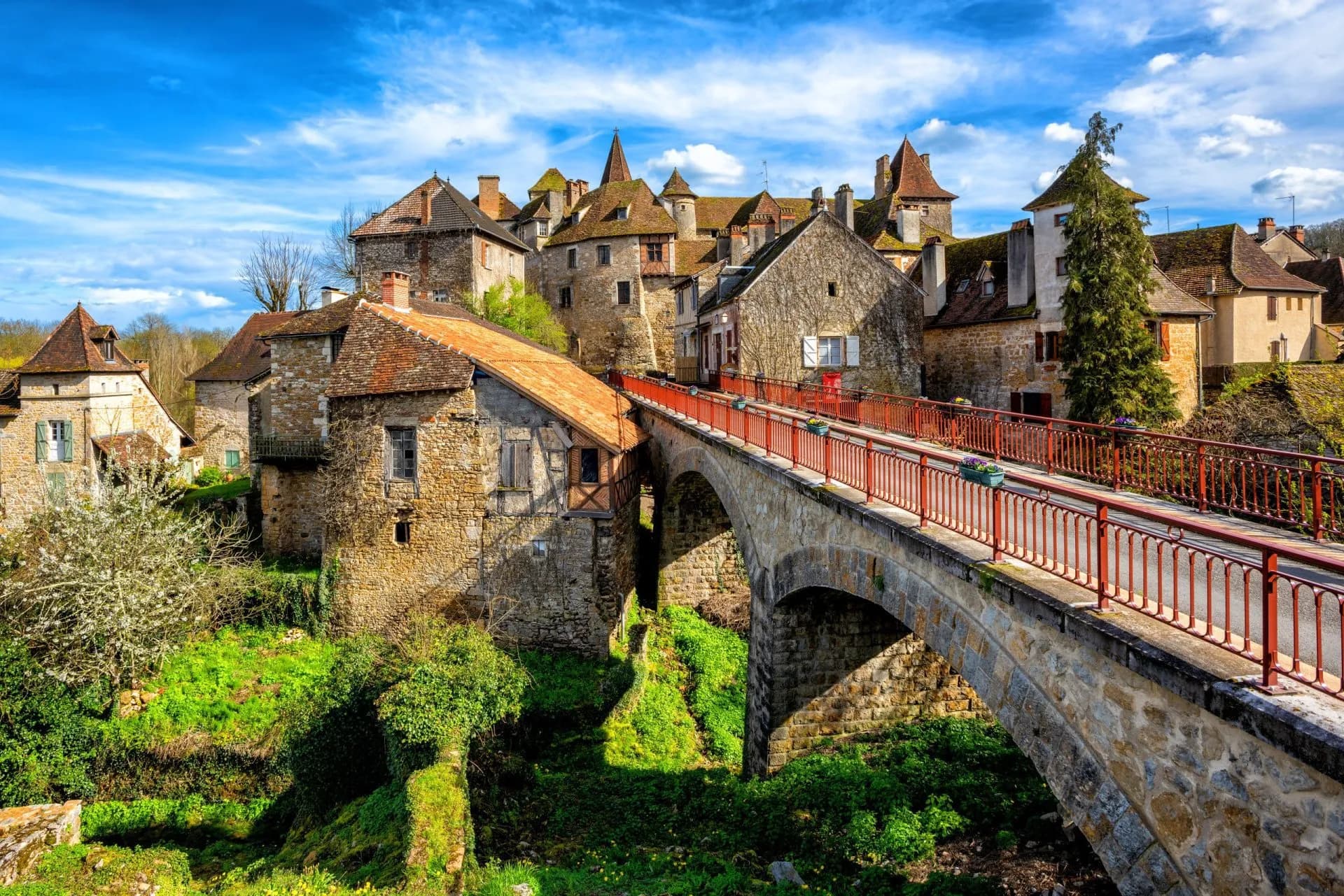 Stone bridge with red railings over lush green ravine leading to historic village buildings.