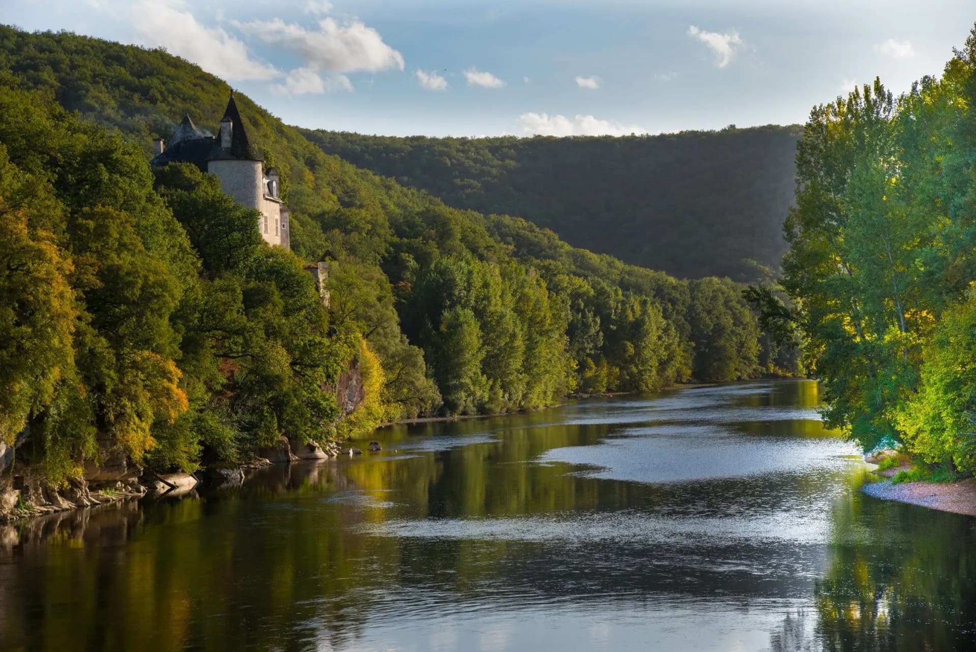 Chateau on forested hillside above a wide river with reflections under a blue sky.