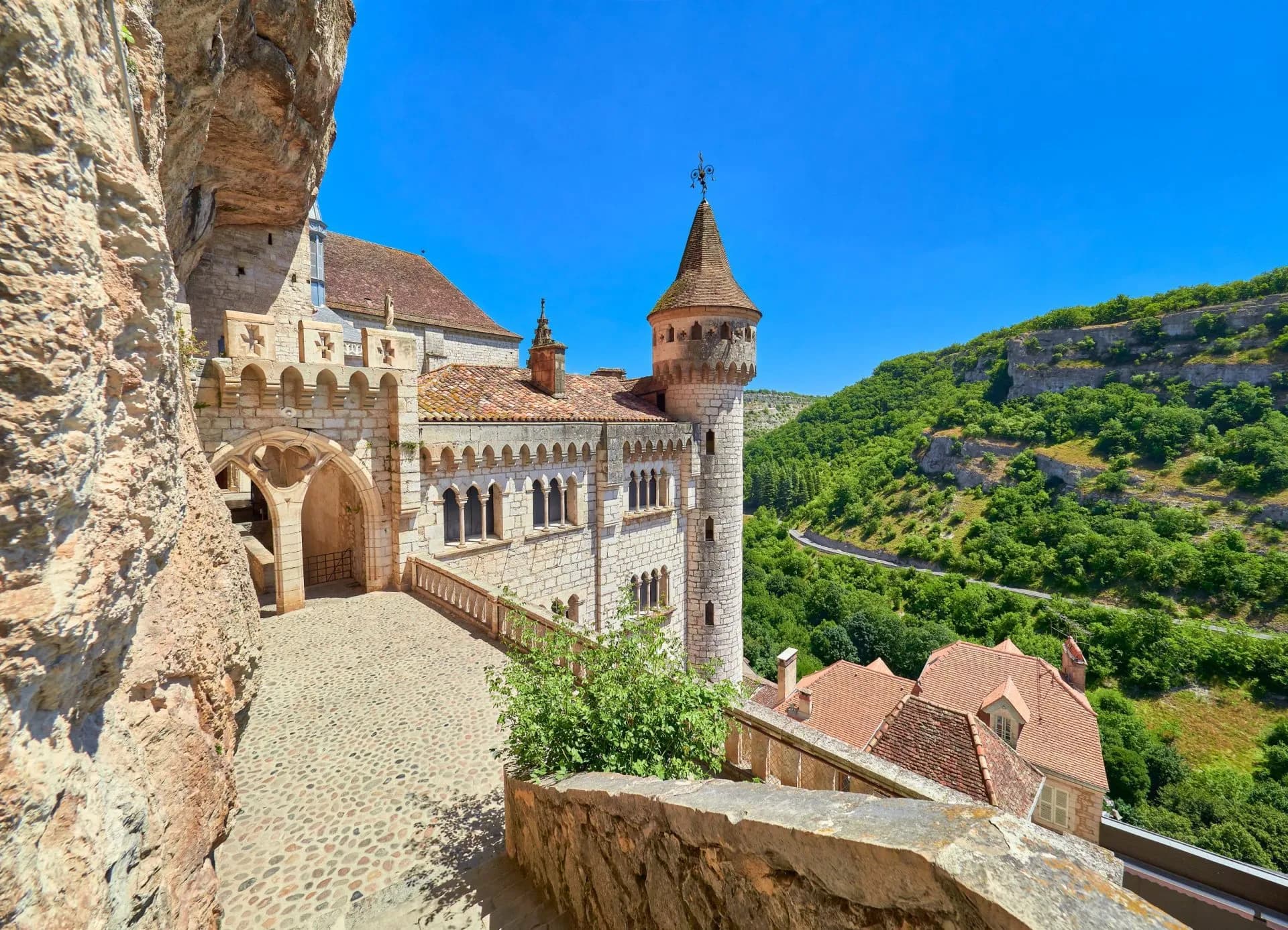 Stone buildings with a turret nestled against a cliff face above a green valley in Rocamadour.