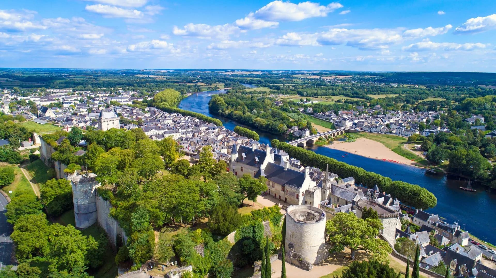 Aerial view of historic town with stone castle ruins beside a wide blue river under a sunny sky.