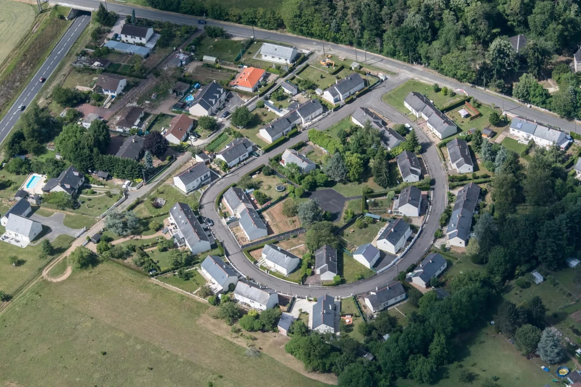 Aerial view of a curved residential street with detached white houses surrounded by green yards and trees near Azay-le-Rid...