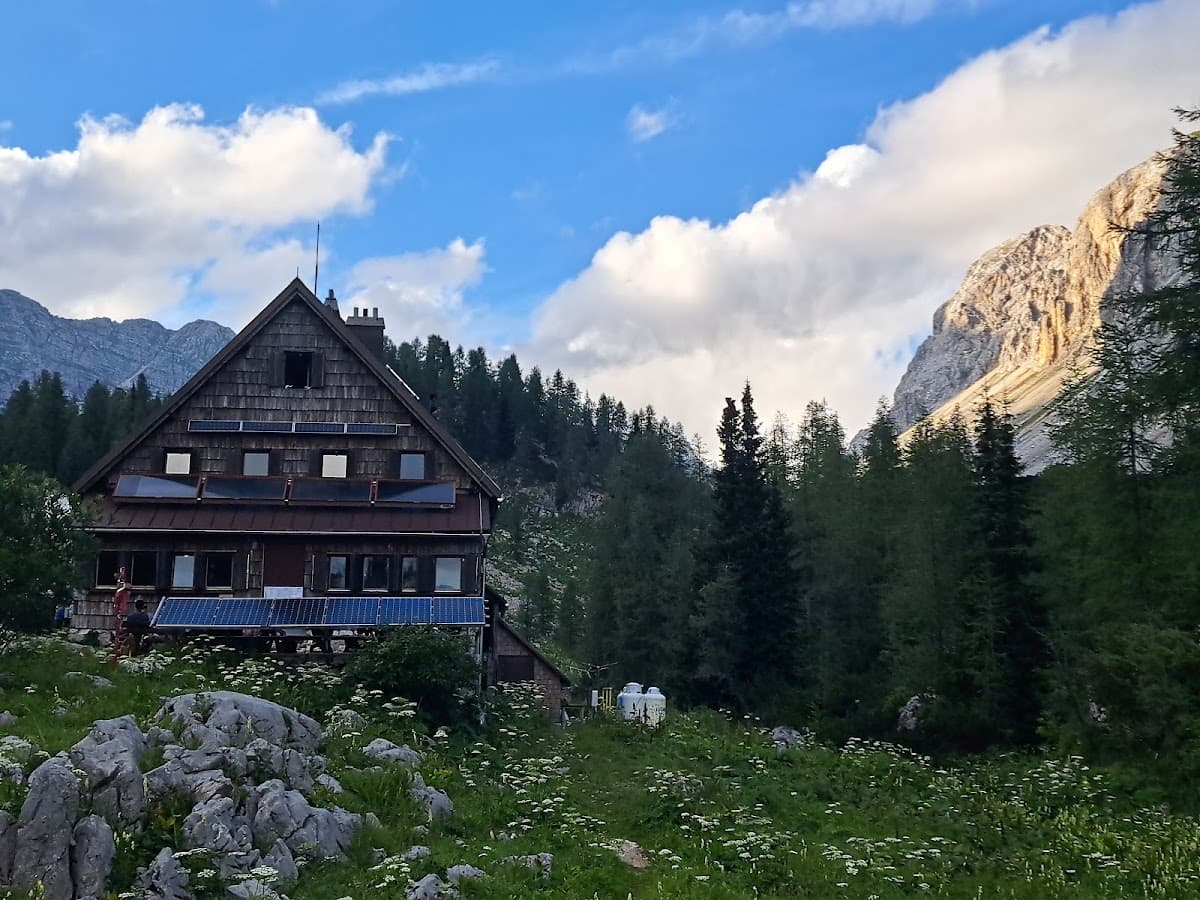Wooden mountain hut with solar panels in a meadow below rocky peaks and pine trees.