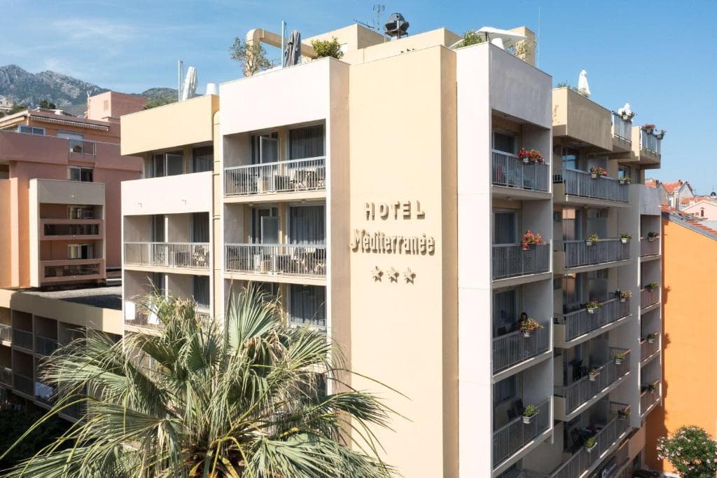 Hotel Mediterranée building with balconies, palm tree, and mountains in the background.