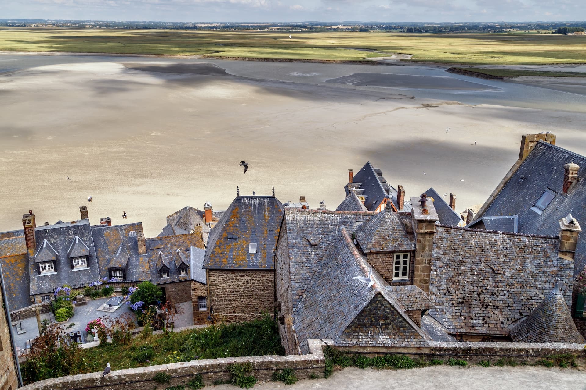 Mont Saint-Michel village rooftops overlooking low tide ocean flats and distant green fields.